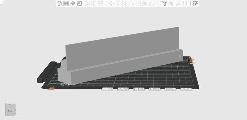May include: A gray rectangular object with a raised back and a lower base, possibly a shelf or display stand. It sits on a black grid-patterned surface, with a black rectangular piece to the left. The design appears to be a 3D model.