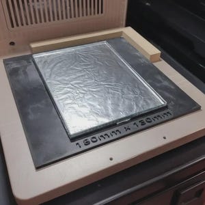 May include: A 3D printer with a beige frame, a black base, and a silver foil-covered platform. The platform is labeled "130mm x 130mm". The printer has a ventilation grill and is on a dark surface.