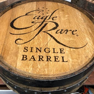 May include: Close-up of a wooden barrel with the words "Eagle Rare SINGLE BARREL" in black lettering. The barrel is made of light brown wood with dark metal bands around the sides. The top of the barrel has a decorative design.