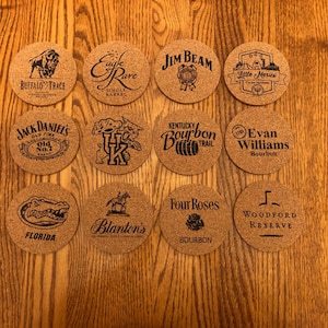 May include: A collection of twelve round cork coasters with various brand logos and text, including "Buffalo Trace," "Jack Daniel's," and "Jim Beam." The coasters are arranged on a wooden surface.