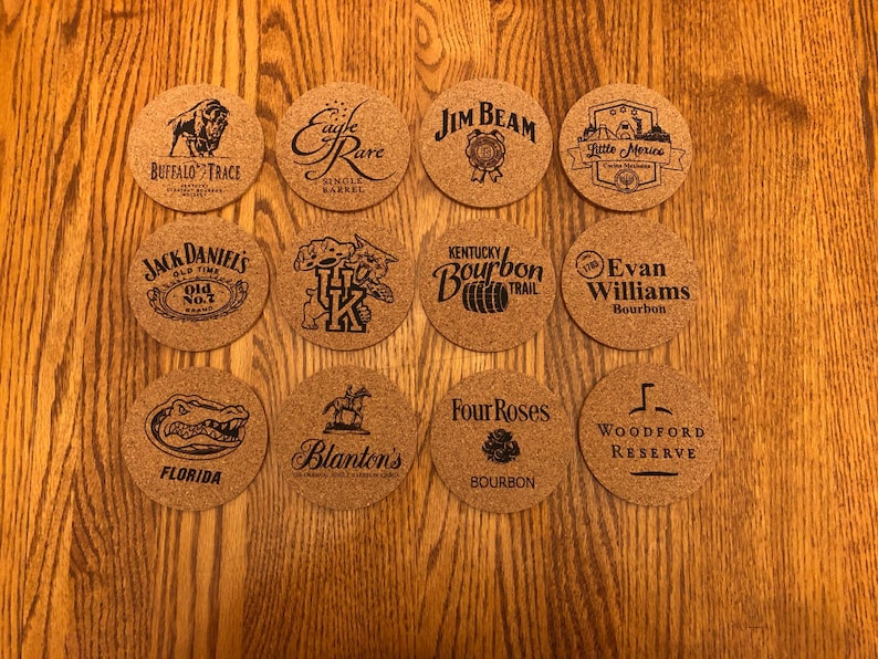 May include: A collection of twelve round cork coasters with various brand logos and text, including "Buffalo Trace," "Jack Daniel's," and "Jim Beam." The coasters are arranged on a wooden surface.