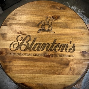Puede incluir: Tablero de mesa redondo de madera con el texto grabado "Blanton's THE ORIGINAL SINGLE BARREL BOURBON" y un logotipo de caballo y jinete. La madera tiene una veta natural y un tono marrón cálido.