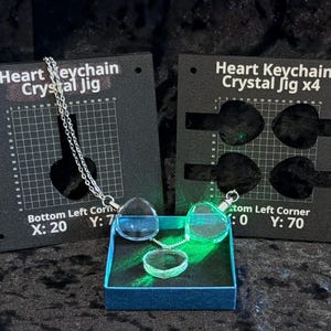 May include: Two black square alignment jigs labeled Heart Keychain Crystal Jig and Heart Keychain Crystal Jig x4 with grid markings. A silver chain connects two clear heart-shaped crystals, with a third crystal resting in a blue box below.
