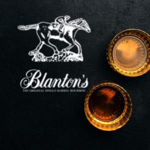 Pode incluir: Uma vista aérea de dois copos de uísque com bordas douradas cheios de líquido âmbar sobre um fundo preto. O logotipo da Blanton's, com um cavalo e um cavaleiro, e o nome da marca são exibidos em branco.
