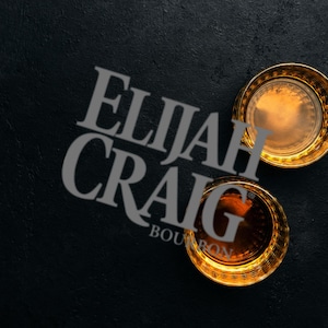 Pode incluir: Dois copos de whisky com bordas douradas cheios de líquido âmbar, sobre um fundo escuro e texturizado. O texto "ELIJAH CRAIG BOURBON" é sobreposto em uma fonte cinza claro, adicionando um toque de sofisticação à composição.