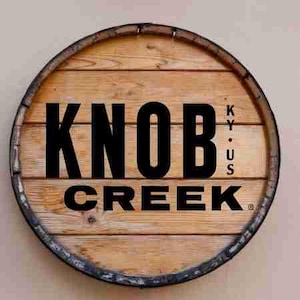 Puede incluir: Letrero redondo de madera con las palabras "KNOB CREEK" en letras negras grandes. El letrero está hecho de tablones de madera y tiene una banda metálica oscura alrededor del borde. Las letras "K.Y.U.S." están apiladas verticalmente a la derecha de "KNOB".