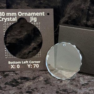 Könnte beinhalten: Eine dunkelbraune quadratische Schablone mit kreisförmigem Ausschnitt und weißem Text 80 mm Ornament Crystal Jig, Bottom Left Corner X: 0 Y: 70, neben einem klaren, runden, facettierten Glasornament mit einem kleinen Loch zum Aufhängen.