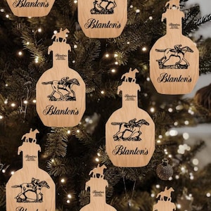 Pode incluir: Enfeites de Natal de madeira em forma de garrafas de uísque, com um desenho de cavalo e cavaleiro e o logotipo "Blanton's". Os enfeites estão pendurados em uma árvore de Natal.