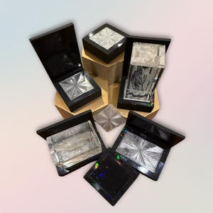Peut inclure: Un assortiment de boîtes carrées noires avec des motifs argentés en forme d'éclat d'étoile. Certaines boîtes sont ouvertes, révélant le motif argenté, tandis que d'autres sont fermées. Une boîte contient une image en noir et blanc. Les boîtes sont disposées sur une surface géométrique dorée.