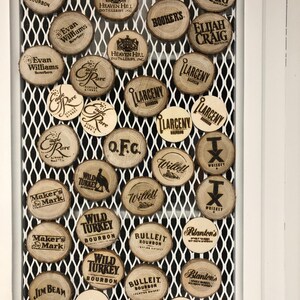 May include: A collection of wooden coasters with various bourbon brand logos, including Four Roses, Jim Beam, and Wild Turkey. The coasters are arranged on a white metal grid, creating a rustic and organized display.