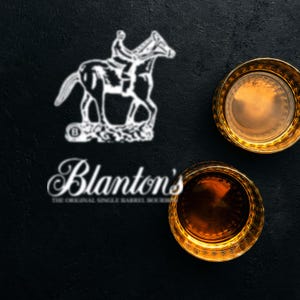 Pode incluir: Dois copos de whisky com bordas douradas cheios de líquido âmbar sobre uma superfície preta. O logotipo da Blanton's, com um cavalo e um cavaleiro, é visível, juntamente com o nome da marca e "THE ORIGINAL SINGLE BARREL BOURBON".