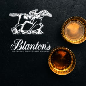 Pode incluir: Dois copos de uísque com bordas douradas cheios de líquido âmbar estão em uma superfície preta. O logotipo da Blanton's, com um cavalo e um cavaleiro, e o nome da marca são exibidos em branco.