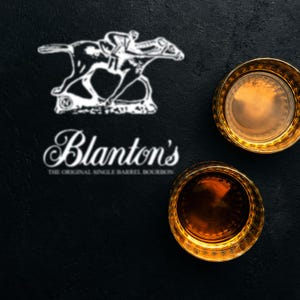 Pode incluir: Dois copos de uísque com bordas douradas cheios de líquido âmbar estão sobre uma superfície preta. O logotipo da Blanton's, com um cavalo e um cavaleiro, e o nome da marca são exibidos acima dos copos.