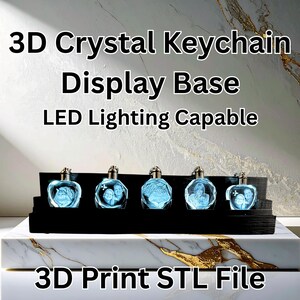 May include: A black display base holding five 3D crystal keychains. Each keychain has a silver cap and features a different image illuminated in blue. The text "3D Crystal Keychain Display Base LED Lighting Capable" is at the top.