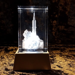 May include: A clear glass sculpture of a rocket launching, encased in a rectangular glass box. The rocket is illuminated, creating a bright, glowing effect. The sculpture sits on a rectangular, light brown base, against a dark, textured background.