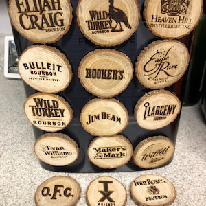 May include: A collection of wooden coasters with various bourbon brand names and logos, including Elijah Craig, Wild Turkey, and Jim Beam. The coasters are round, made from wood slices, and feature engraved text.