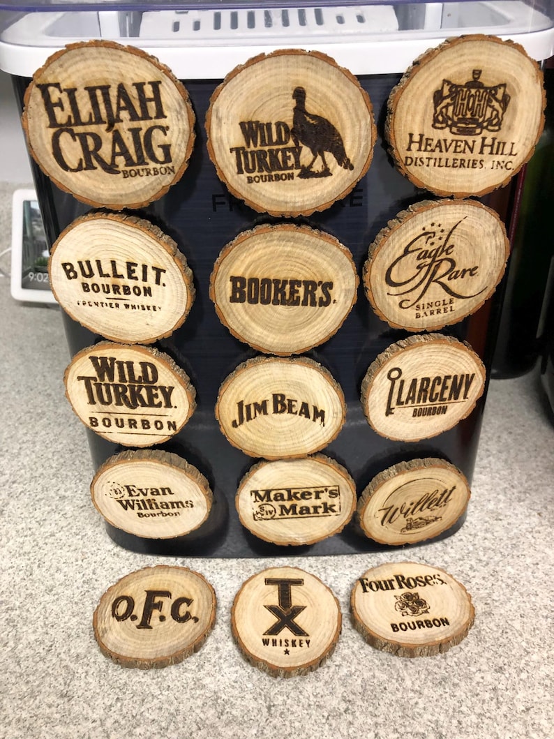 May include: A collection of wooden coasters with various bourbon brand names and logos, including Elijah Craig, Wild Turkey, and Jim Beam. The coasters are round, made from wood slices, and feature engraved text.