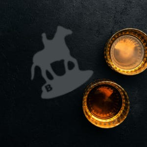 Pode incluir: Dois copos de whisky com bordas douradas cheios de líquido âmbar estão sobre uma superfície escura. À esquerda, uma silhueta cinza de um cavalo e cavaleiro, com a letra "B" na parte inferior. Os copos são provavelmente para servir bebidas espirituosas.