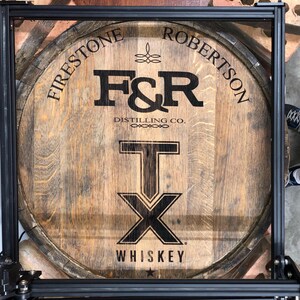 Pode incluir: Uma cabeça de barril de uísque de madeira com o texto "Firestone Robertson" e "F&R Distilling Co." em preto. A cabeça do barril também apresenta um grande design "T" sobre "X", com a palavra "Whiskey" abaixo.