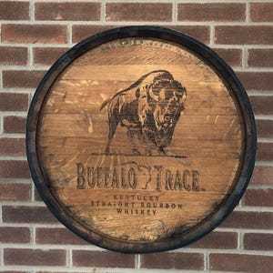 May include: Circular wooden sign with a dark rim, featuring a bison illustration and the words "Buffalo Trace Kentucky Straight Bourbon Whiskey." The sign is mounted on a brick wall.