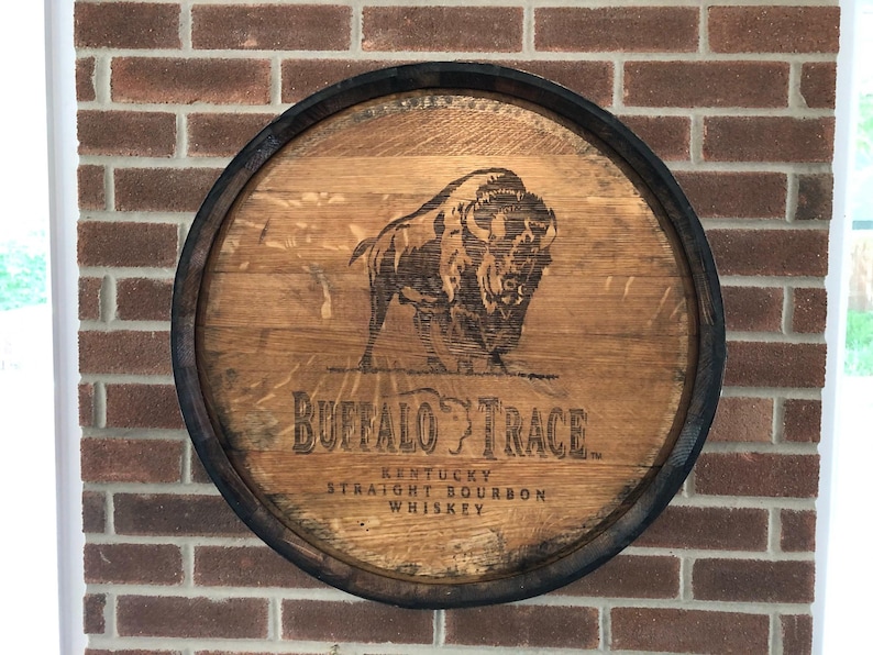May include: Round wooden sign with a dark rim, featuring a bison illustration and the words "Buffalo Trace Kentucky Straight Bourbon Whiskey." The sign is mounted on a brick wall.