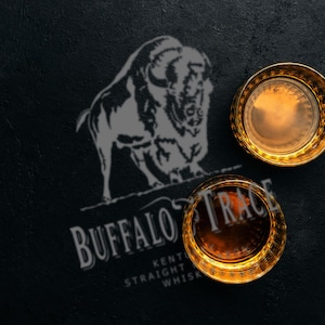 May include: Two gold-rimmed whiskey glasses filled with amber liquid sit on a dark surface. The Buffalo Trace logo and a bison silhouette are visible. The text reads "Kentucky Straight Whiskey."