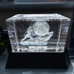 May include: A clear, rectangular crystal block with an illuminated top, displaying a 3D image of a sphere and abstract shapes. The base is black, and the text "BELIEVE DOUBLE-DIGIT GROWTH" is visible.