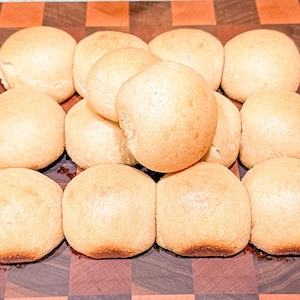 Könnte beinhalten: Eine Ansammlung frisch gebackener, goldbrauner Brötchen, die auf einer karierten Holzoberfläche angeordnet sind. Die Brötchen haben eine weiche, runde Form und eine leicht dunklere Kruste am Boden, was darauf hindeutet, dass sie warm und essfertig sind.