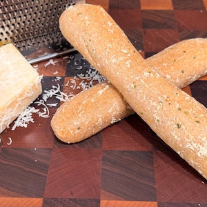 May include: Two golden-brown breadsticks are crossed on a wooden cutting board. A block of cheese and a cheese grater are in the background. The breadsticks have a sprinkle of herbs and a dusting of flour.