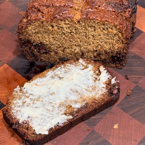 Può includere: Una pagnotta di pane appena sfornato con una fetta davanti, spalmata di burro. Il pane ha una crosta marrone scuro e un interno marrone chiaro. La fetta di pane è su un tagliere di legno.