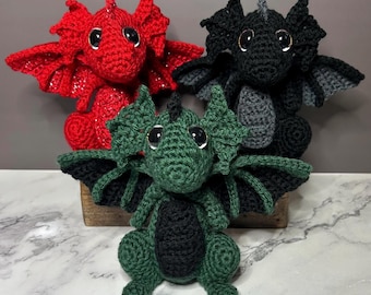 Puede incluir: Tres dragones de peluche tejidos a ganchillo con grandes ojos negros brillantes y alas texturizadas. Un dragón es rojo brillante, otro es negro sólido con detalles grises, y el dragón frontal es verde oscuro con vientre y alas negras.