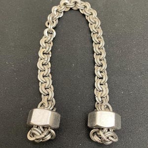 May include: A silver-colored chain bracelet with a unique design. The bracelet features a series of interlocking links forming a flexible chain. The ends of the bracelet are capped with hexagonal metal pieces.