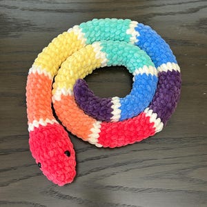 Könnte beinhalten: Ein regenbogenfarbenes Häkel-Schlangenspielzeug. Die Schlange ist zu einem Kreis zusammengerollt, mit dem Kopf zum Betrachter. Die Schlange ist aus weichem, flauschigem Garn gefertigt und hat ein helles, farbenfrohes Design.