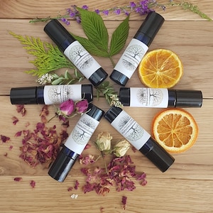 May include: Six glass roller bottles with black caps and white labels. The labels feature a tree logo and text that reads "Recovering With Nature" and "Happy Vibes". The bottles are arranged in a circle on a wooden surface with dried flowers and citrus fruit slices.