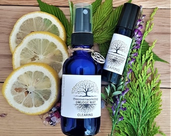 May include: Two blue glass bottles with white labels. The bottles are labeled "The Coven Workshop Smudge Mist Clearing" and "The Coven Workshop Body Smudge Clearing". The bottles are surrounded by lemon slices, green leaves, and purple flowers.