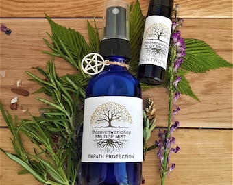 May include: Two blue glass bottles with white labels. The larger bottle is a spray bottle with the text "thecovenworkshop SMUDGE MIST EMPATH PROTECTION". The smaller bottle is a roll-on bottle with the text "thecovenworkshop BODY SMUDGE PATH PROTECTION".