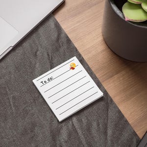 May include: A white notepad with black lines and the words "To do:" printed at the top. A small cartoon image of a person with yellow hair and a red scarf is in the top right corner. The notepad is on a dark grey surface.