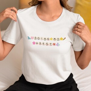 May include: White t-shirt with the phrase "DOING MY BEST" in colorful letters and symbols. The shirt has a crew neck and short sleeves. The text includes a heart, flower, star, and pencil.