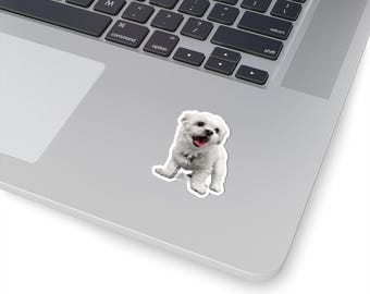May include: A white sticker featuring a small, fluffy dog with its mouth open and tongue visible. The sticker is placed on a silver laptop, with the keyboard partially visible. The dog appears to be a Maltese or similar breed.