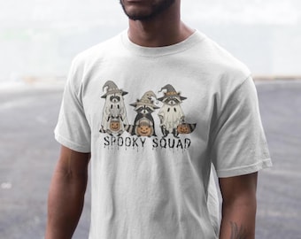Op de afbeelding: Een wit T-shirt met korte mouwen en een afbeelding van drie wasberen in Halloween-kostuums, inclusief heksenhoeden en spooklakens, die elk een kleine oranje pompoen vasthouden. De tekst SPOOKY SQUAD staat eronder gedrukt.
