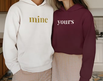 Puede incluir: Dos sudaderas con capucha a juego, una color crema con "mine" en letras doradas y la otra burdeos con "yours" en blanco. Ambas tienen un bolsillo delantero y una capucha. La pareja lleva pantalones cortos a cuadros a juego.