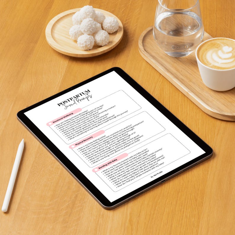 May include: A digital tablet displaying "Postpartum Journal Prompts" with prompts for emotional wellbeing, physical recovery, and bonding with baby. A white stylus rests beside the tablet on a wooden surface, along with a plate of coconut macaroons, a glass of water, and a cup of coffee.
