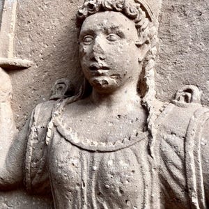 Könnte beinhalten: Ein verwittertes Steinrelief, das eine Figur mit geflochtenem Haar darstellt. Sie trägt ein strukturiertes Gewand mit Schulterträgern und hält ein Schwert aufrecht in der rechten Hand. Die Oberfläche zeigt Altersspuren mit körniger Textur.