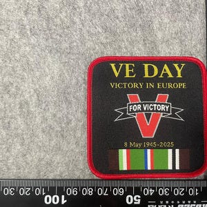 Peut inclure: Un écusson brodé noir et rouge avec le texte "VE DAY VICTORY IN EUROPE" et un "V" rouge avec le texte "FOR VICTORY" au-dessus. L'écusson comporte également un ruban avec des rayures rouges, blanches, bleues et vertes et le texte "8 May 1945-2025".