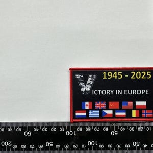 May include: A red, white, and blue embroidered patch with the text "1945-2025 Victory in Europe" and a stylized "V" in the center. The patch features flags from various countries, including Canada, the United Kingdom, the United States, and France.