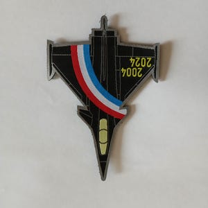 Può includere: Una toppa ricamata nera e grigia di un aereo da combattimento con i colori della bandiera francese sul lato. Il testo "2007" e "2027" è sulla toppa.