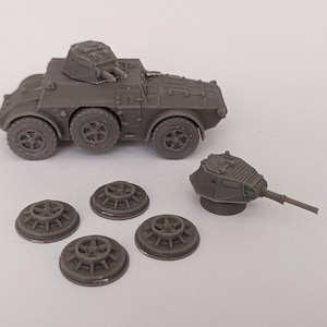 May include: A gray plastic model of a military armored car with a turret on top. The car has four wheels and is sitting on a white surface. There are five gray plastic model wheels and a gray plastic model turret on the surface.
