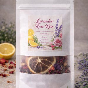 May include: A white pouch with a clear window displaying dried botanicals. The label reads "Lavender Rose Kiss" with illustrations of lemon slices, lavender, and a rose. A half lemon, strawberries, and lavender sprigs are also visible.