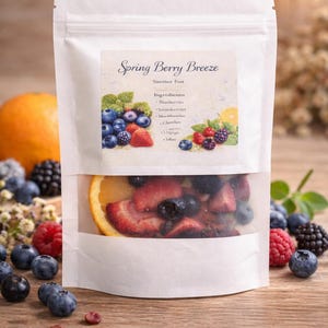 May include: A white pouch with a clear window, filled with sliced strawberries, blueberries, and a lemon slice. The label reads "Spring Berry Breeze" with a list of ingredients. Fresh berries are scattered around the pouch on a wooden surface.