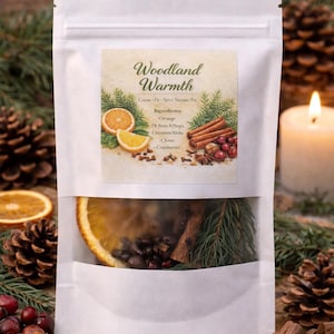 May include: A white pouch with a clear window, filled with a spice blend. The label reads "Woodland Warmth" and lists ingredients like orange slices, cinnamon sticks, and cranberries. The background includes pine cones and a lit candle.